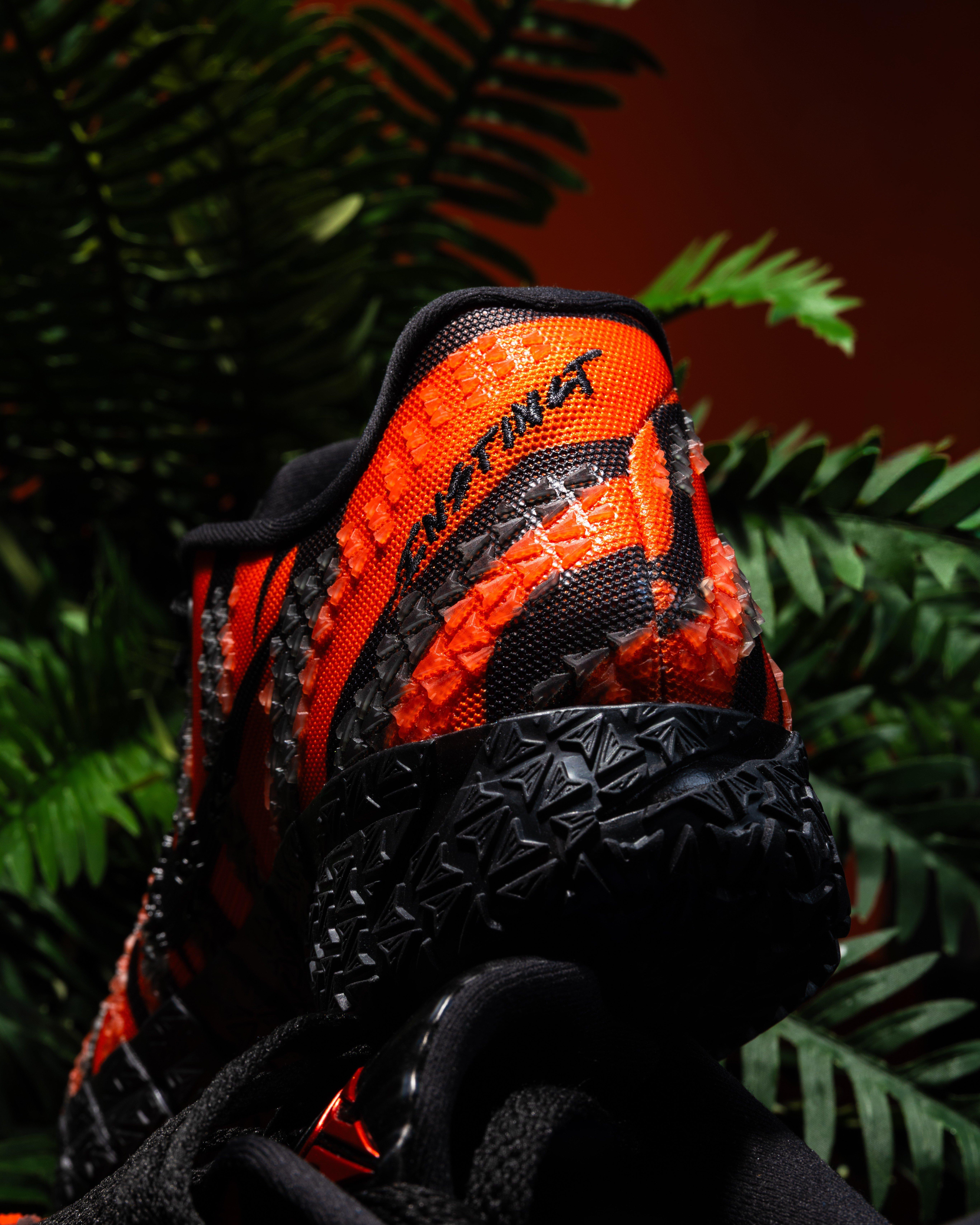 Rear view of a Nike Ja 3 sneaker with bright orange and black textured upper, visible &ldquo;12AM&rdquo; text on the heel, and black outsole, surrounded by green leaves.