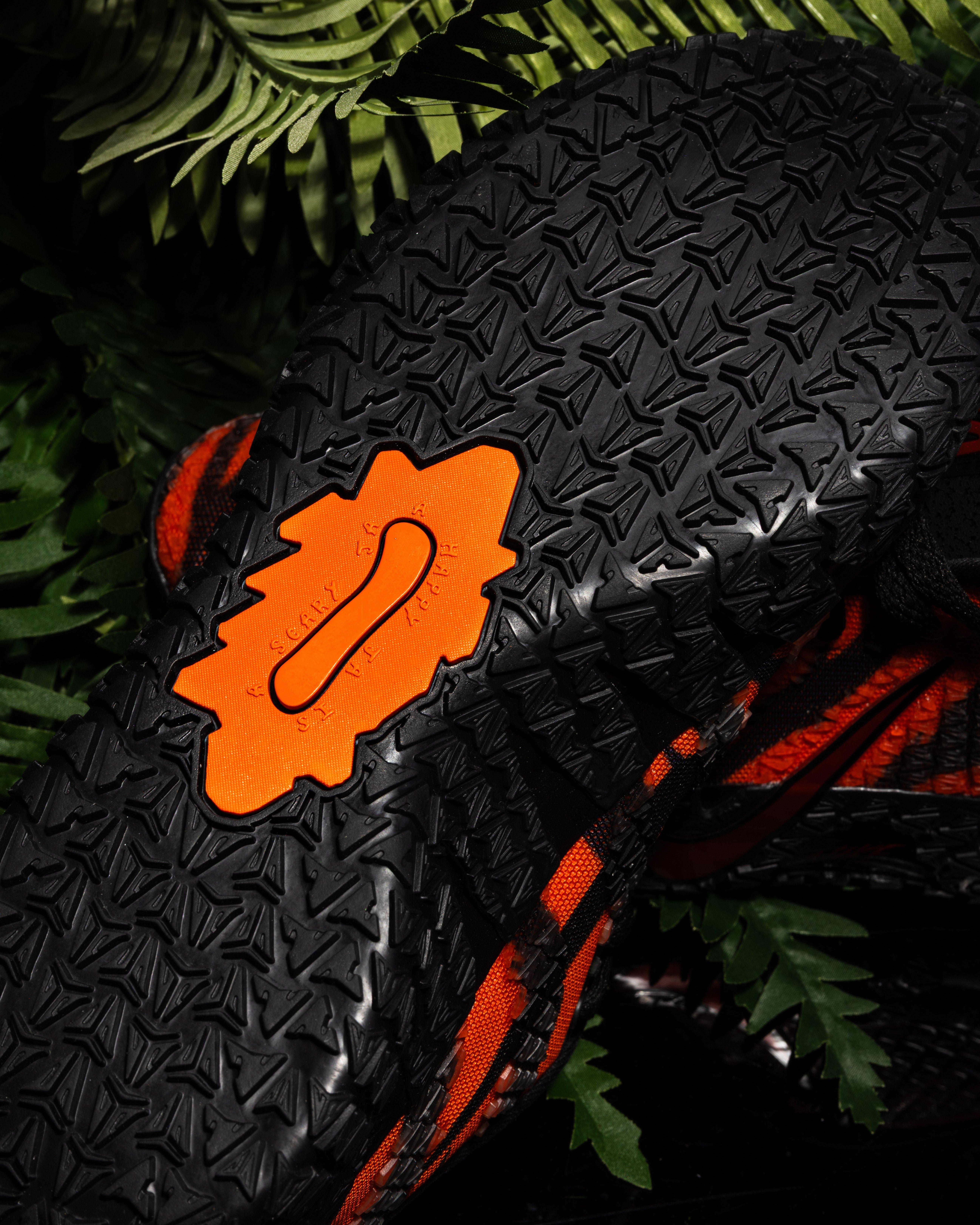 Close-up of the outsole of a Nike Ja 3 sneaker showing a black traction pattern with an orange midfoot plate, set against green foliage.