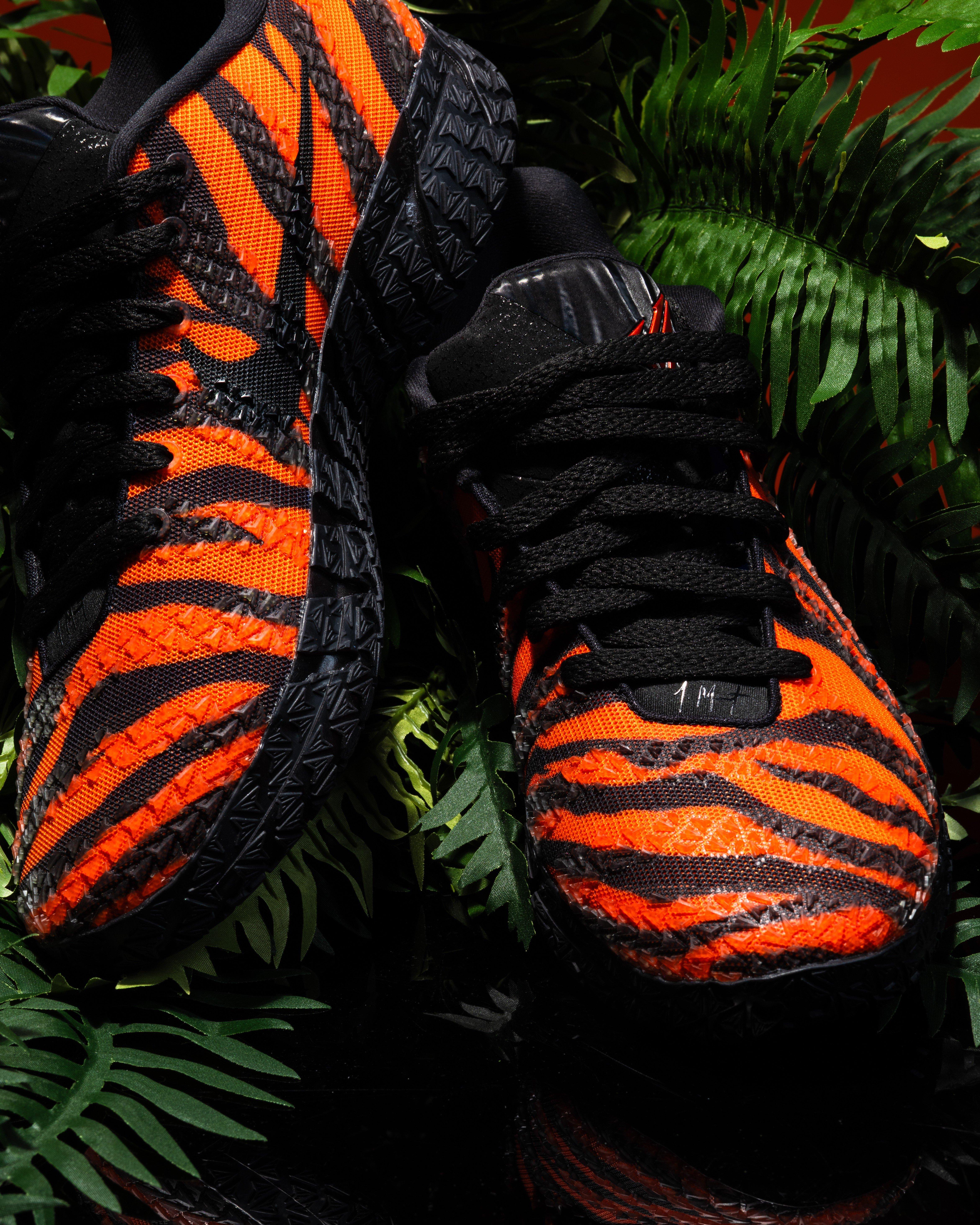 Top-down view of a Nike Ja 3 sneaker highlighting black laces over an orange and black tiger-striped upper, with textured detailing and green foliage background.