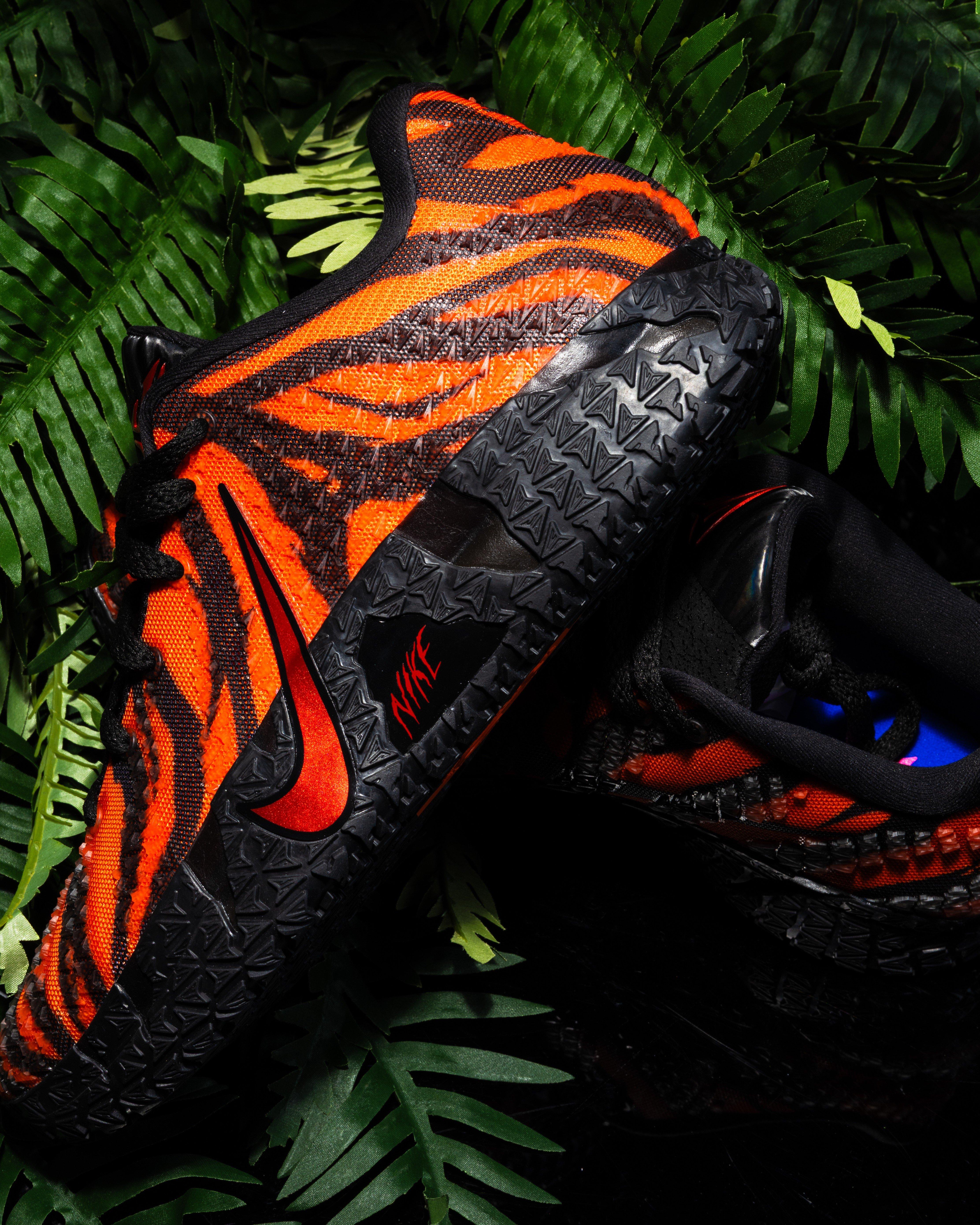 Side view of a Nike Ja 3 sneaker featuring an orange and black tiger-striped upper with a black Nike Swoosh and rugged black sole, displayed among green tropical leaves.