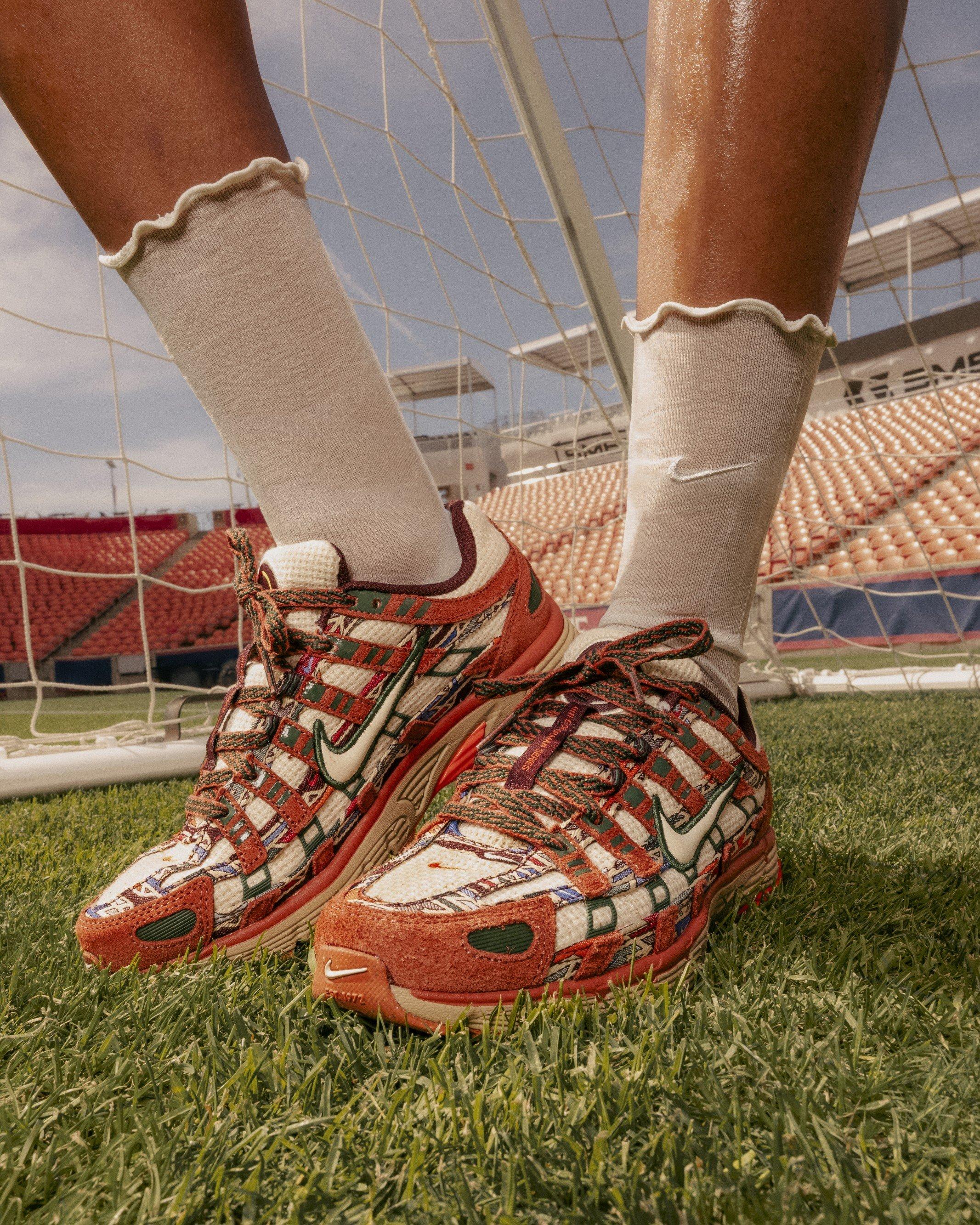 Close-up shot of a person's feet on a green soccer pitch wearing the Nike P-6000 N7 sneakers. The shoes feature an intricate multi-colored textile pattern inspired by Native weaving traditions, accented by dusty peach suede overlays, a white Nike swoosh, and patterned green laces, paired with cream-colored ruffled crew socks.