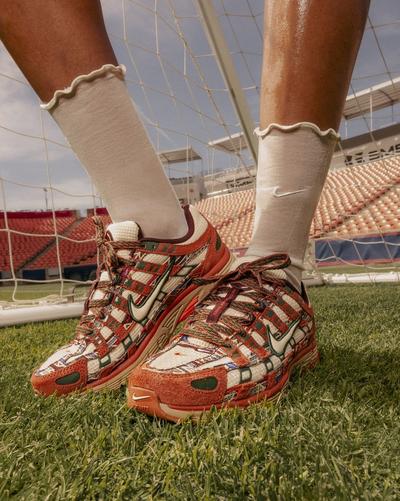 Close-up of patterned Nike sneakers with rust, cream and green details, styled with cream Nike socks on a soccer field in front of a goal net.