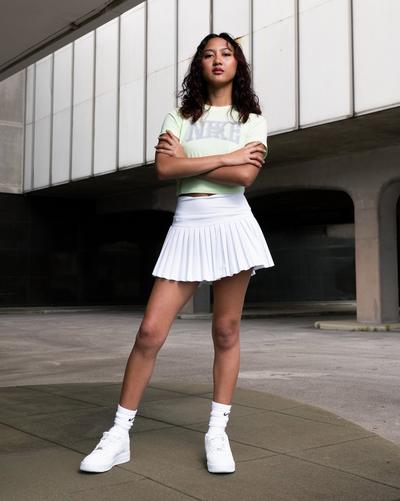 Woman in Nike Women's Sportswear Slim Crop Tee, Gold Hinge Women's Pleated Tennis Skort - White, Nike Socks, and Nike Air Force 1 '07 LE "White/White" Women's Shoe