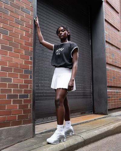 A woman wears a Nike Sportswear Collection Knit Tank Top, Gold Hinge Pleated Tennis Skort, and Nike Shox R4 White-Metallic Silver sneakers while leaning against a brick wall.