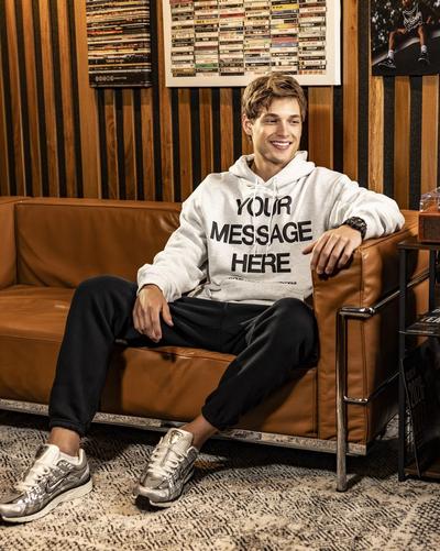 Person seated on a brown leather couch wearing a gray Nike hoodie with &ldquo;Your Message Here&rdquo; text, black pants and silver Nike sneakers in a music-themed room.