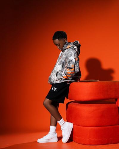 Child model leaning against an orange chair wearing a gray and orange patterned Off-White hoodie, black shorts, white socks and white Nike sneakers against an orange background.