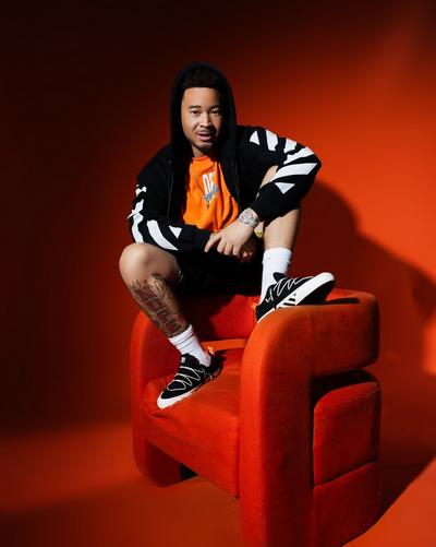 Model seated on an orange chair wearing a black hoodie with white sleeve graphics, orange T-shirt, black shorts, white socks and black Off-White sneakers against an orange background.