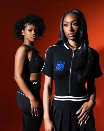Two models standing against an orange background wearing black Off-White apparel, including a zip-up top, cropped top, leggings and skirt with white and blue logo details.