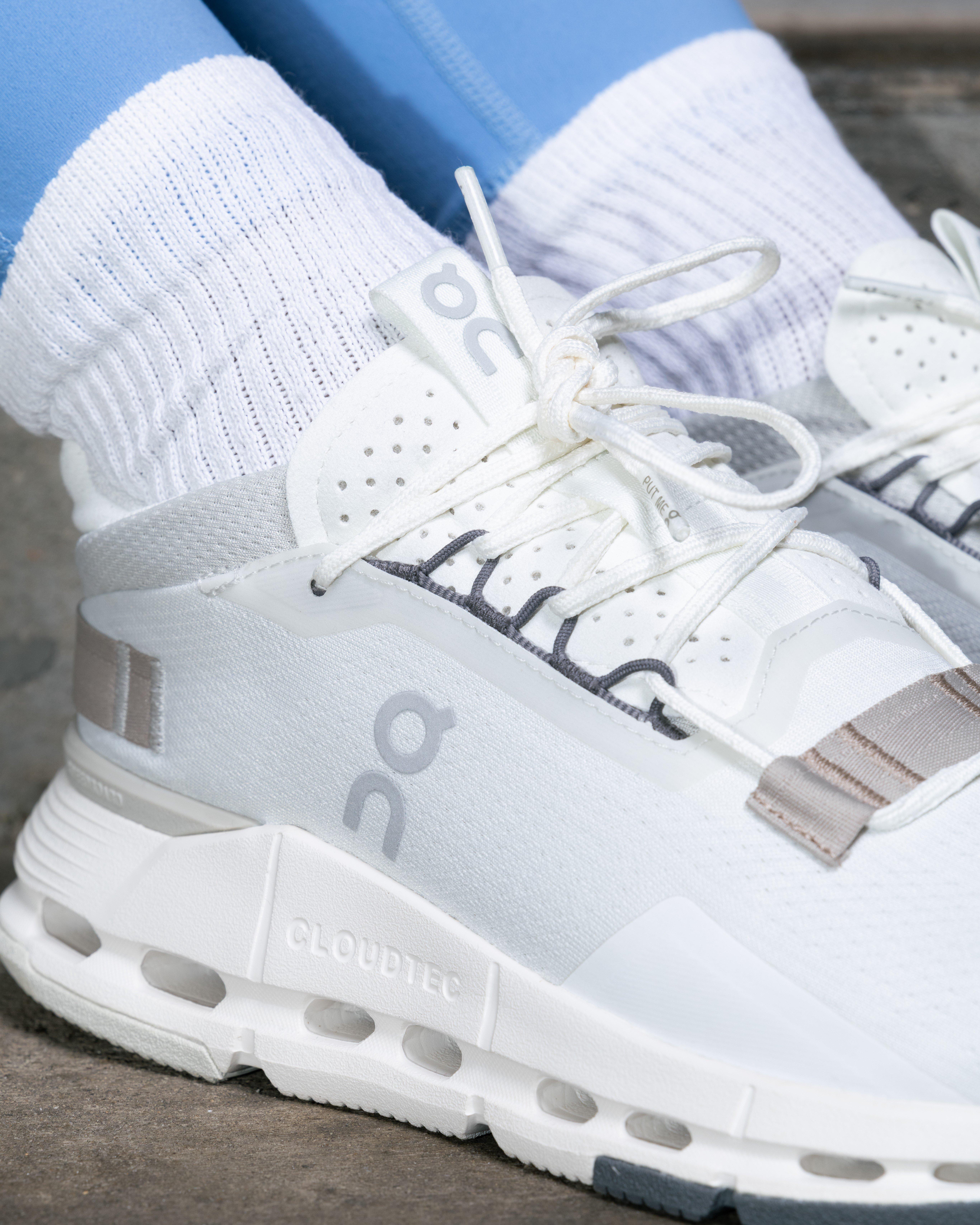 A close-up shot of the white laces and upper part of an On sneaker. The shoes are white and have the "On" logo on the side. A person's legs, wearing light blue leggings and white ribbed socks, are also visible.