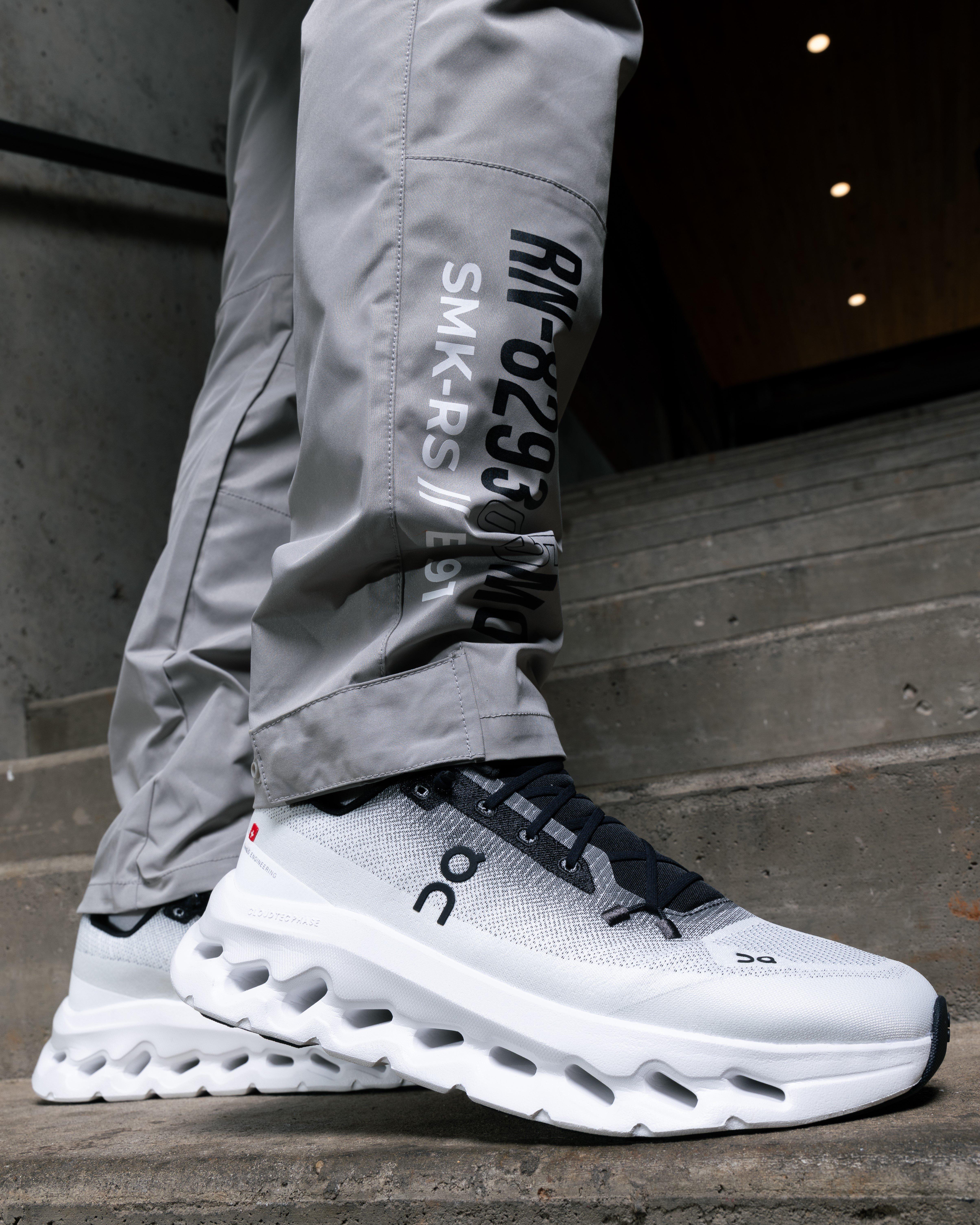 A close-up, low-angle shot of a person's legs walking down concrete steps. They are wearing light gray pants with black text on the leg, and white and black On running shoes with a thick, textured white sole.