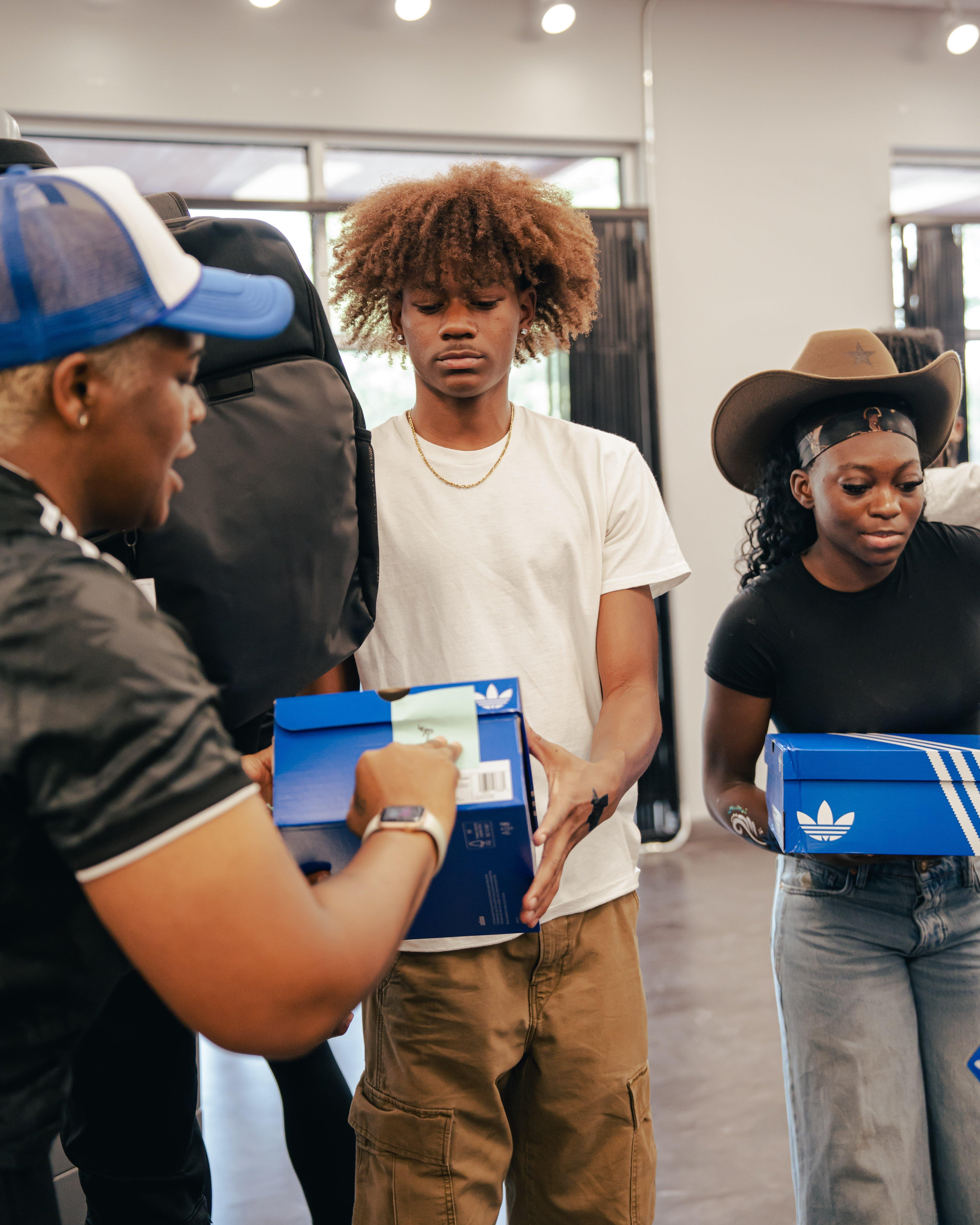 Two young people, a man and a woman, are being given blue shoe boxes. The man in the center has a brown afro and is wearing a white t-shirt and khaki pants. The woman on the right wears a cowboy hat and a black top. The person on the left is handing the man a box.