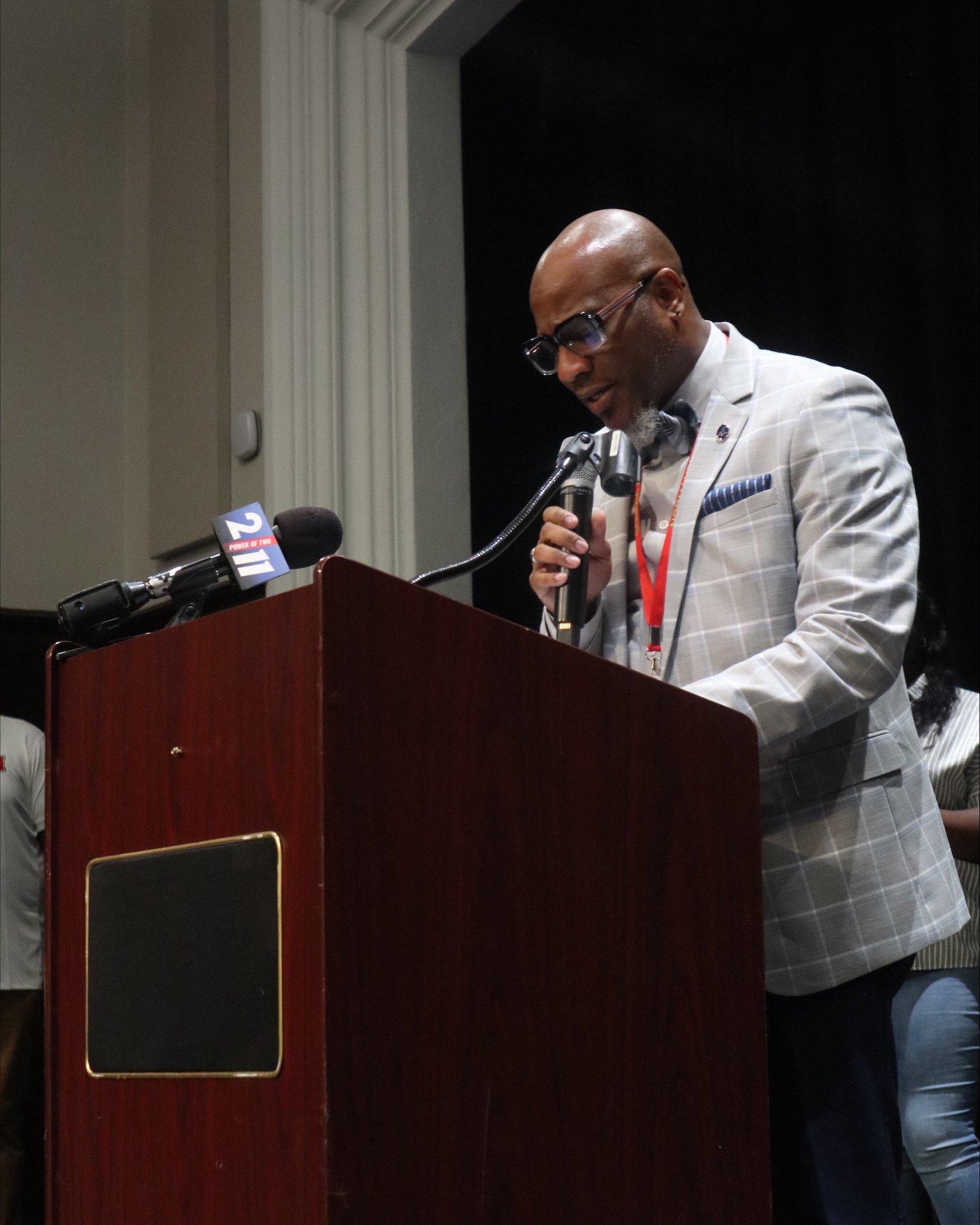 A speaker in a grey plaid suit addressing the audience from a wooden podium during the First String presentation event at Normandy High School.