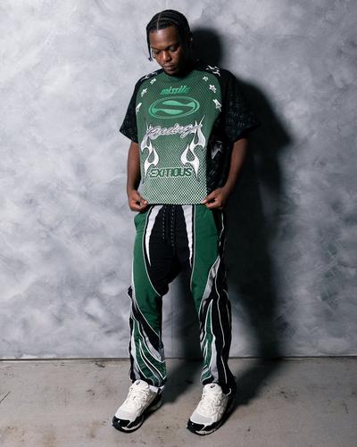Model wearing a green, black and white Pursue moto graphic shirt with matching racing-style pants and white ASICS sneakers against a gray studio wall.