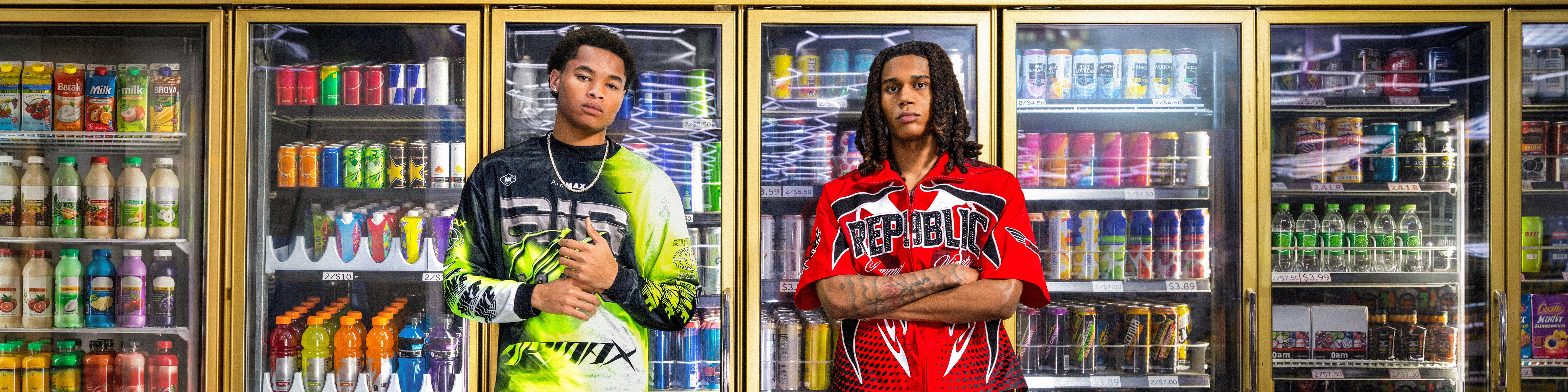 Group of six young adults standing together on a rooftop at night, wearing trendy streetwear and athletic-inspired outfits with bold prints, numbers, and camo patterns, city lights visible in the background.