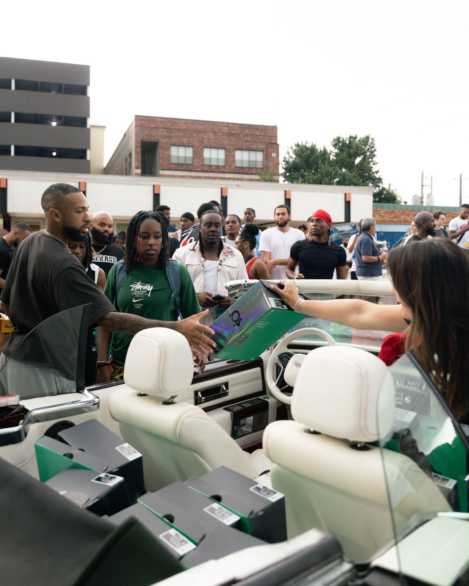 Community event participant receiving a Nike KD shoebox from a convertible filled with giveaway sneakers as attendees gather outdoors.