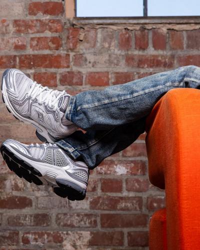 Close-up of silver and white Saucony running shoes styled with acid-wash jeans while seated on an orange chair against a brick wall.