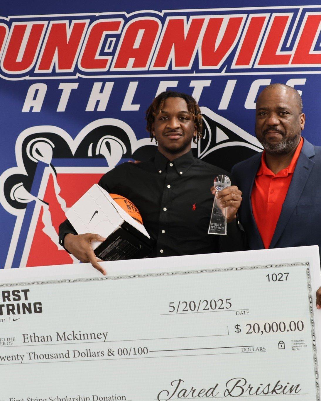  A young man and an older man stand side-by-side, smiling, in front of a large, oversized check. Both have dark skin. The young man on the left wears a black collared shirt with a small red polo emblem and has dreadlocks. He holds a white box with a Nike swoosh and the words "FIRST STRING" in orange, and a clear trophy. The older man on the right wears a navy blue suit jacket over an orange collared shirt. The oversized check is white with "FIRST STRING" printed prominently on the left. It is dated "5/20/2025" and is made out to "Ethan Mckinney" for "$20,000.00." In the background, a red, white, and blue banner with "DUNCANVILLE ATHLETICS" and a panther logo is visible.