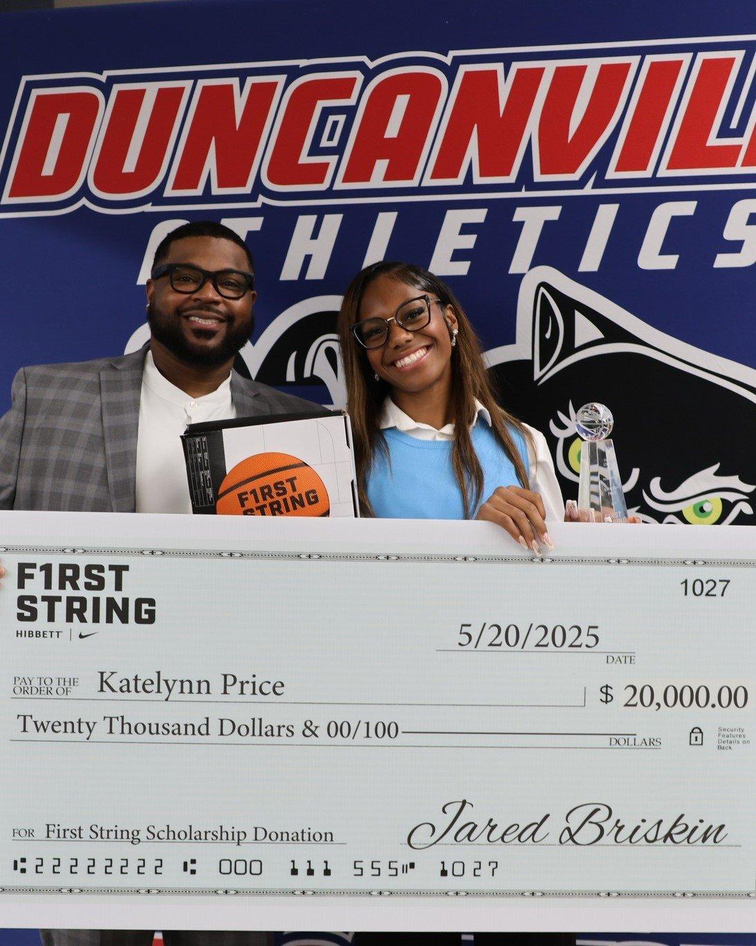  A man and a young woman, both with dark skin and wearing glasses, stand smiling behind a large, oversized check in a brightly lit indoor setting. The man on the left wears a grey suit jacket over a white collared shirt and holds a black box or plaque that says "FIRST STRING" in orange. The young woman on the right wears a light blue sleeveless top over a white collared shirt and holds a trophy. The check is white with "FIRST STRING" printed prominently on the left and is dated "5/20/2025," made out to "Katelynn Price" for "$20,000.00." In the background, a large banner or wall features "DUNCANVILLE ATHLETICS" in red and blue lettering, along with an athletic panther logo