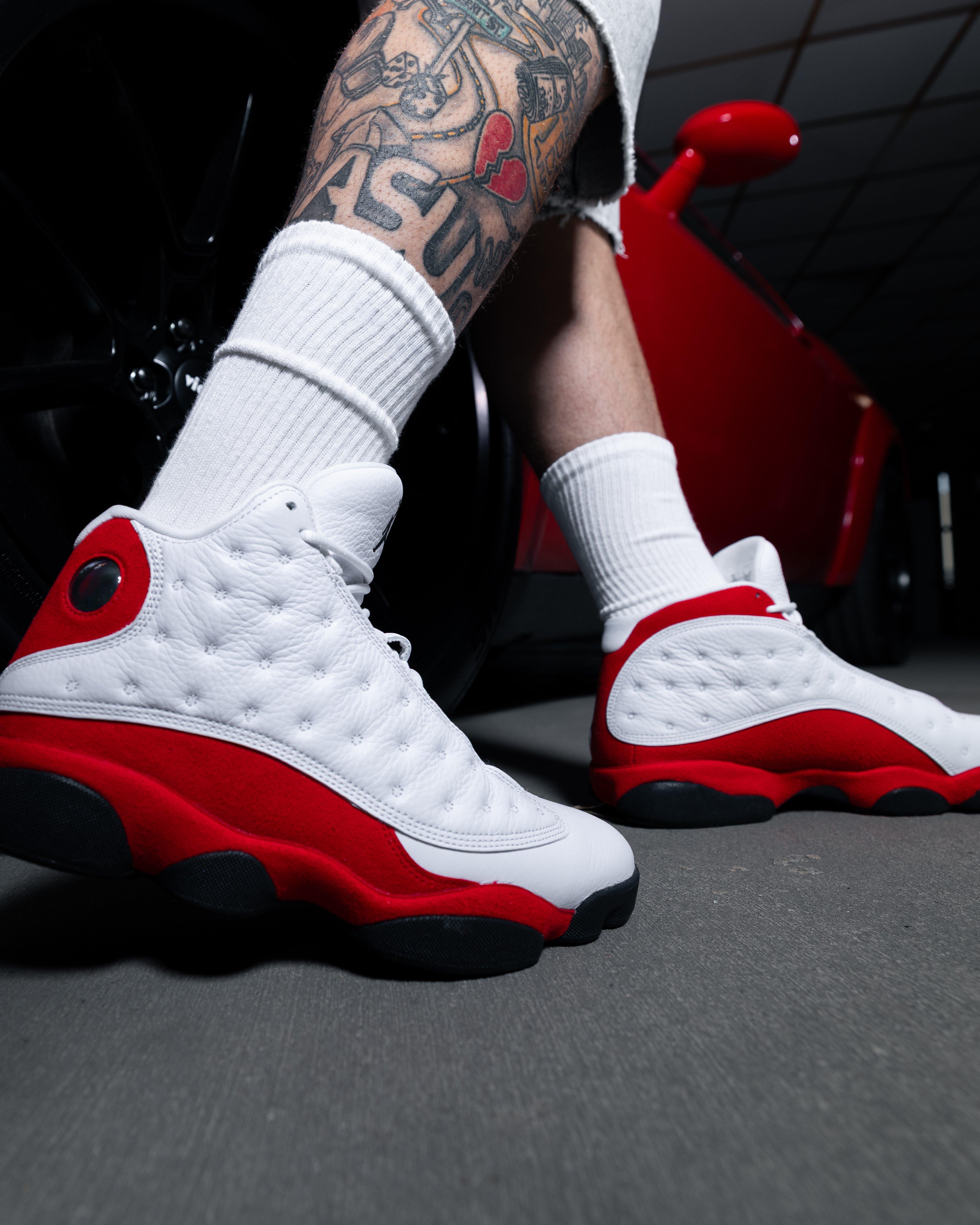 A close-up view of a person wearing white and red athletic sneakers with black soles, standing beside a red car. The person is dressed in white socks and has a tattooed lower leg visible. The photo is taken from a low angle, emphasizing the details and texture of the shoes.