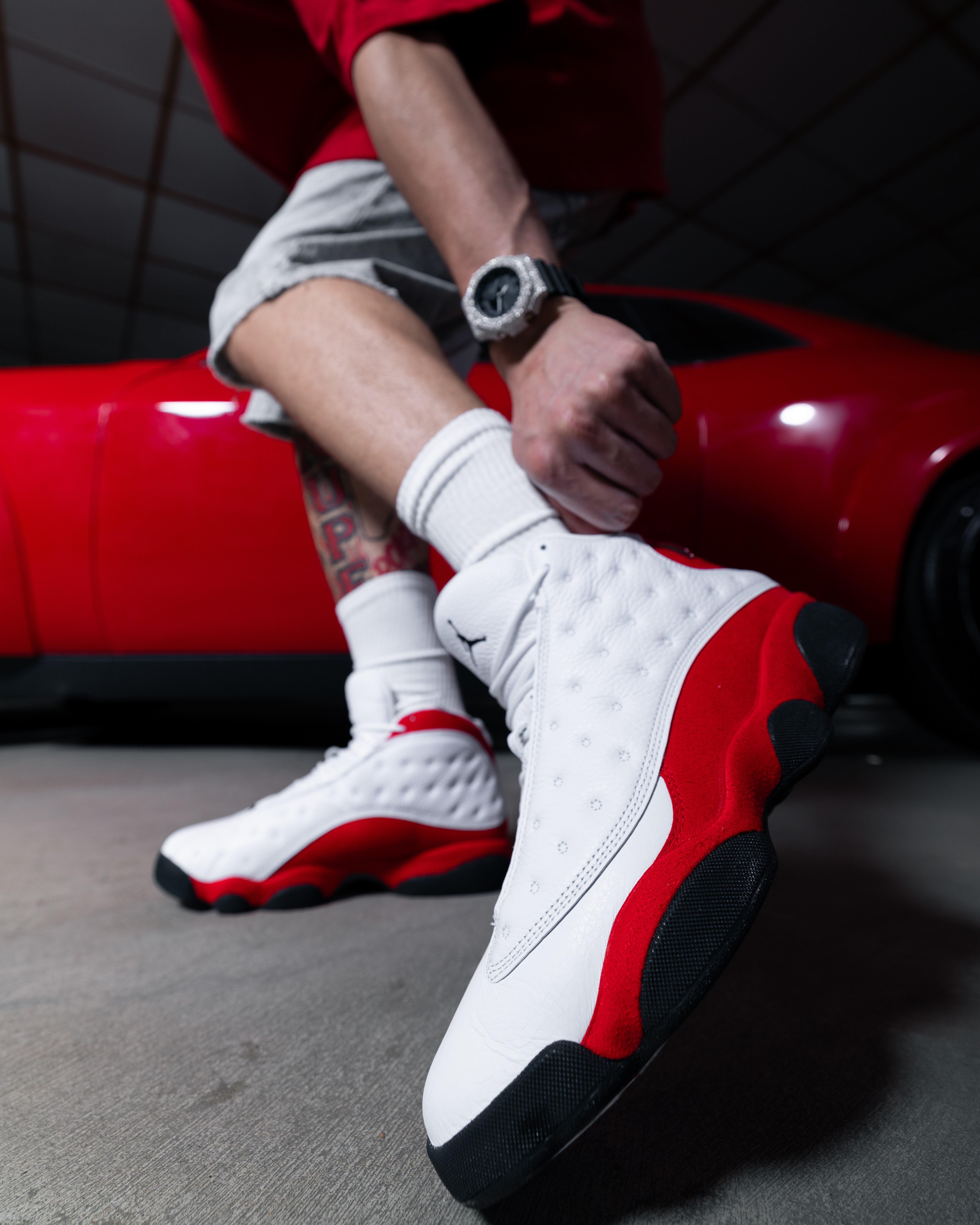 A person wearing white and red athletic sneakers with black soles is bending down slightly, adjusting one shoe. The person is beside a red car, wearing white socks, rolled-up light shorts, and a wristwatch. The low-angle view highlights the shoes&rsquo; design and the car in the background.