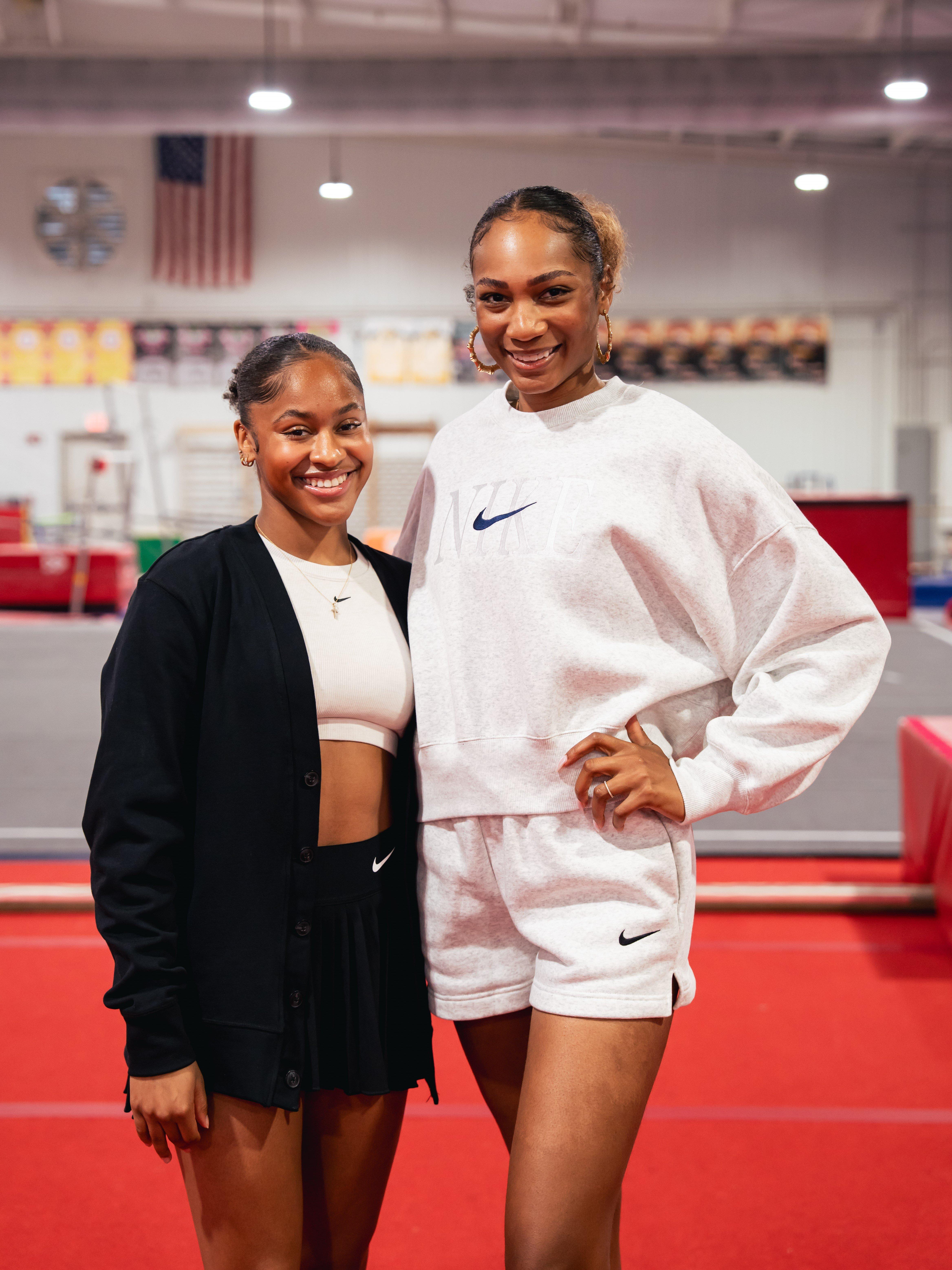 A full-body shot of two women standing side by side. On the left, a smiling Skye Blakely is wearing a black cardigan over a white crop top and black pleated skirt. On the right, another woman is wearing a light gray Nike sweatshirt and matching shorts.