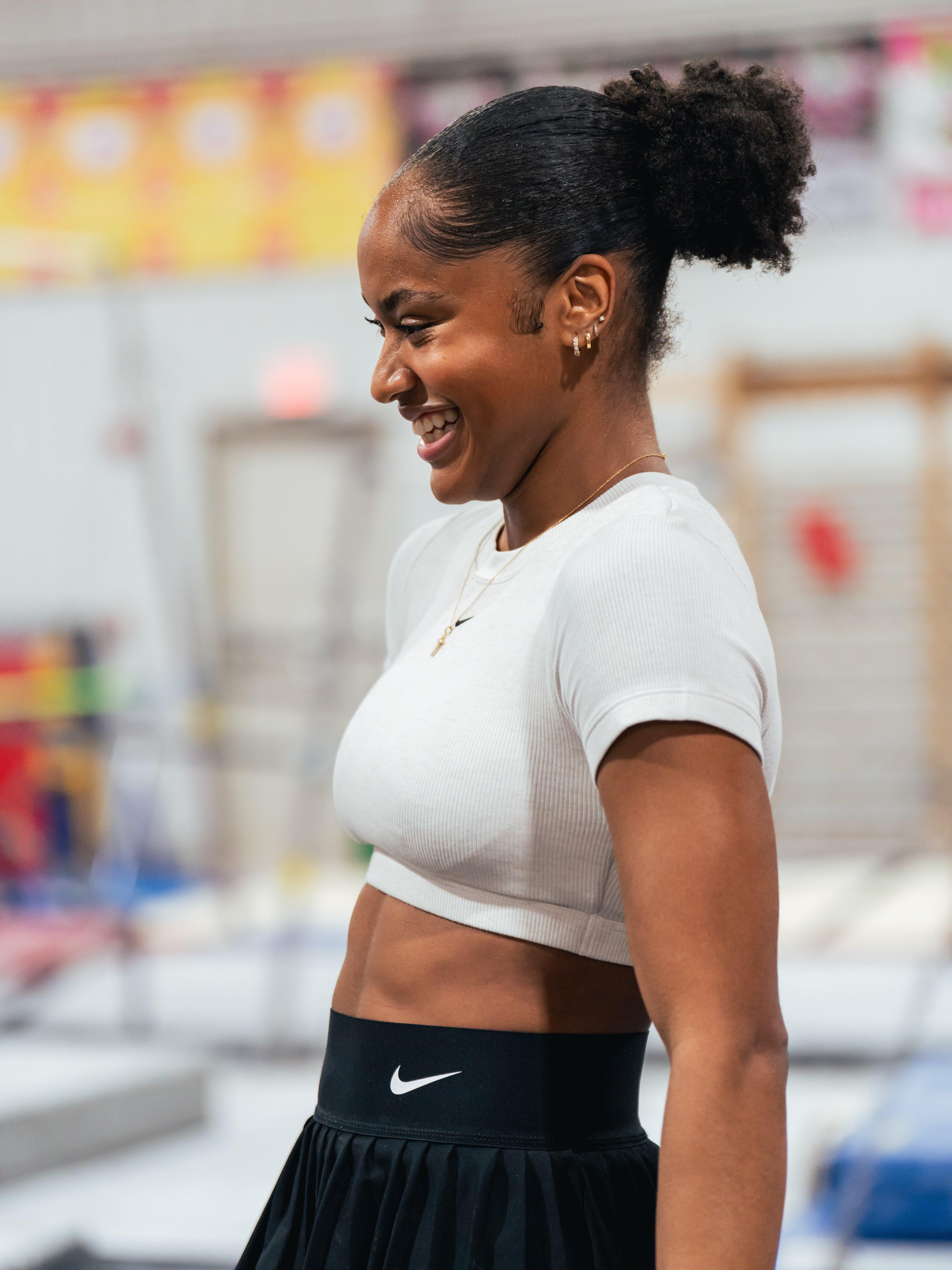  A profile shot of Skye Blakely smiling. Her hair is in a high ponytail. She is wearing a white ribbed crop top and a black pleated skirt with a Nike logo on the waistband.