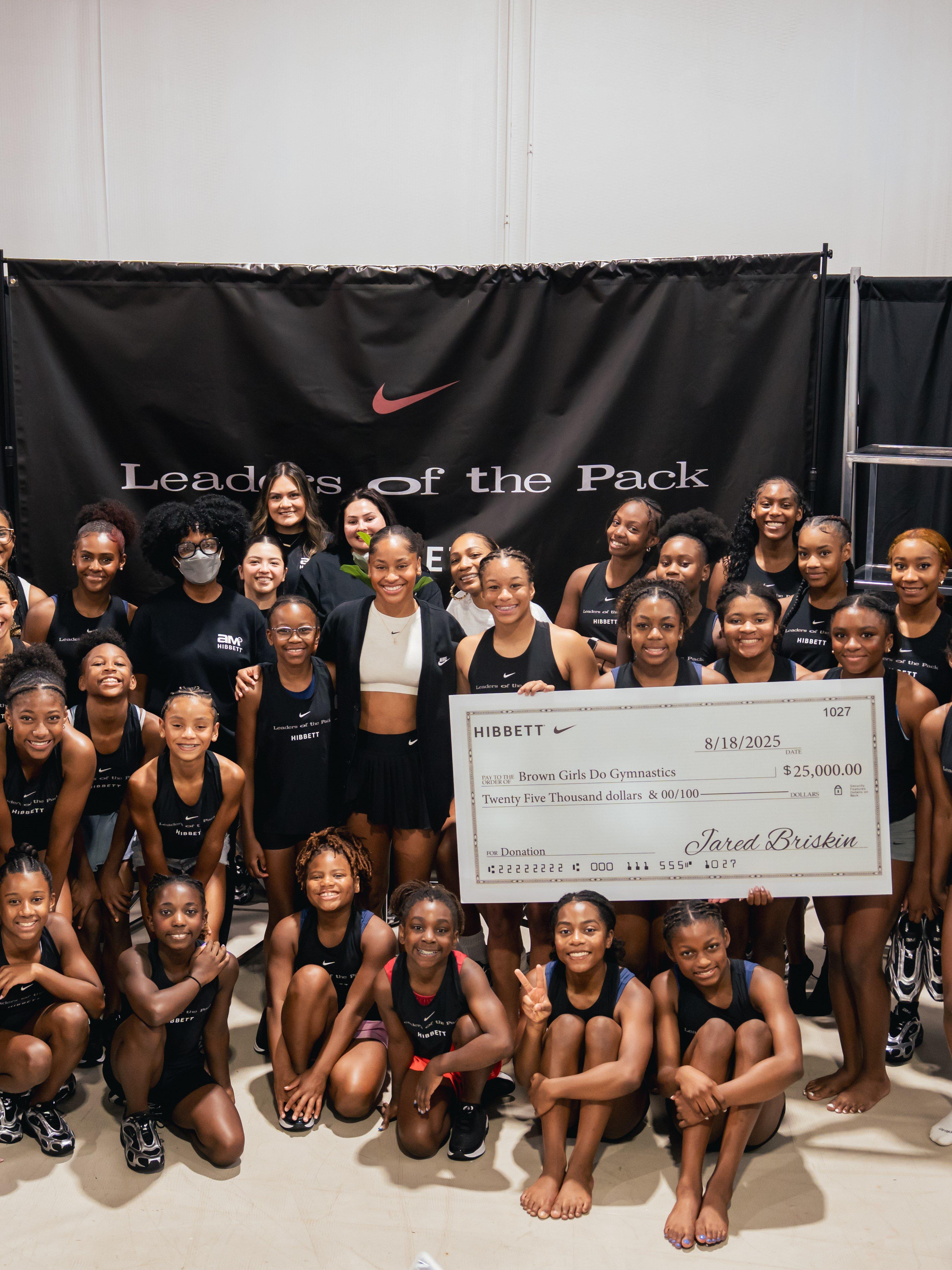 A group of young female gymnasts and adult staff pose with a giant check for $25,000. Skye Blakely is in the center holding the check. A black banner behind them reads "Leaders of the Pack" and "Nike."