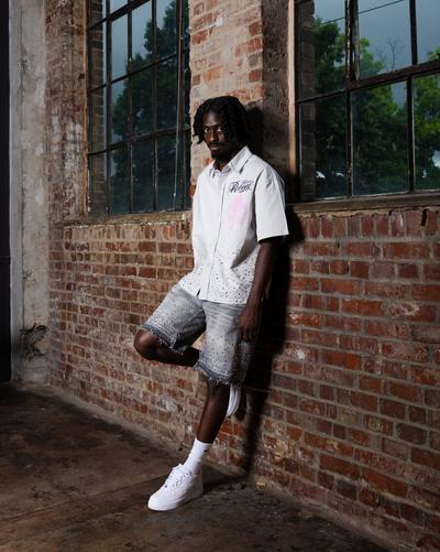 Model wearing a white short-sleeve button-up shirt, embellished denim shorts and white sneakers while standing against a brick wall with large windows.