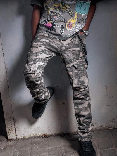 Close-up of a person wearing a gray motorsport graphic T-shirt, gray camouflage cargo pants with multiple pockets and black sneakers against a concrete wall.