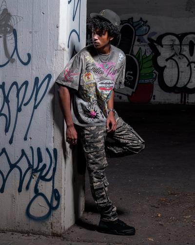 Person wearing a gray motorsport graphic T-shirt, gray camouflage cargo pants, a black baseball cap and black sneakers while posing beside a graffiti-covered concrete wall.