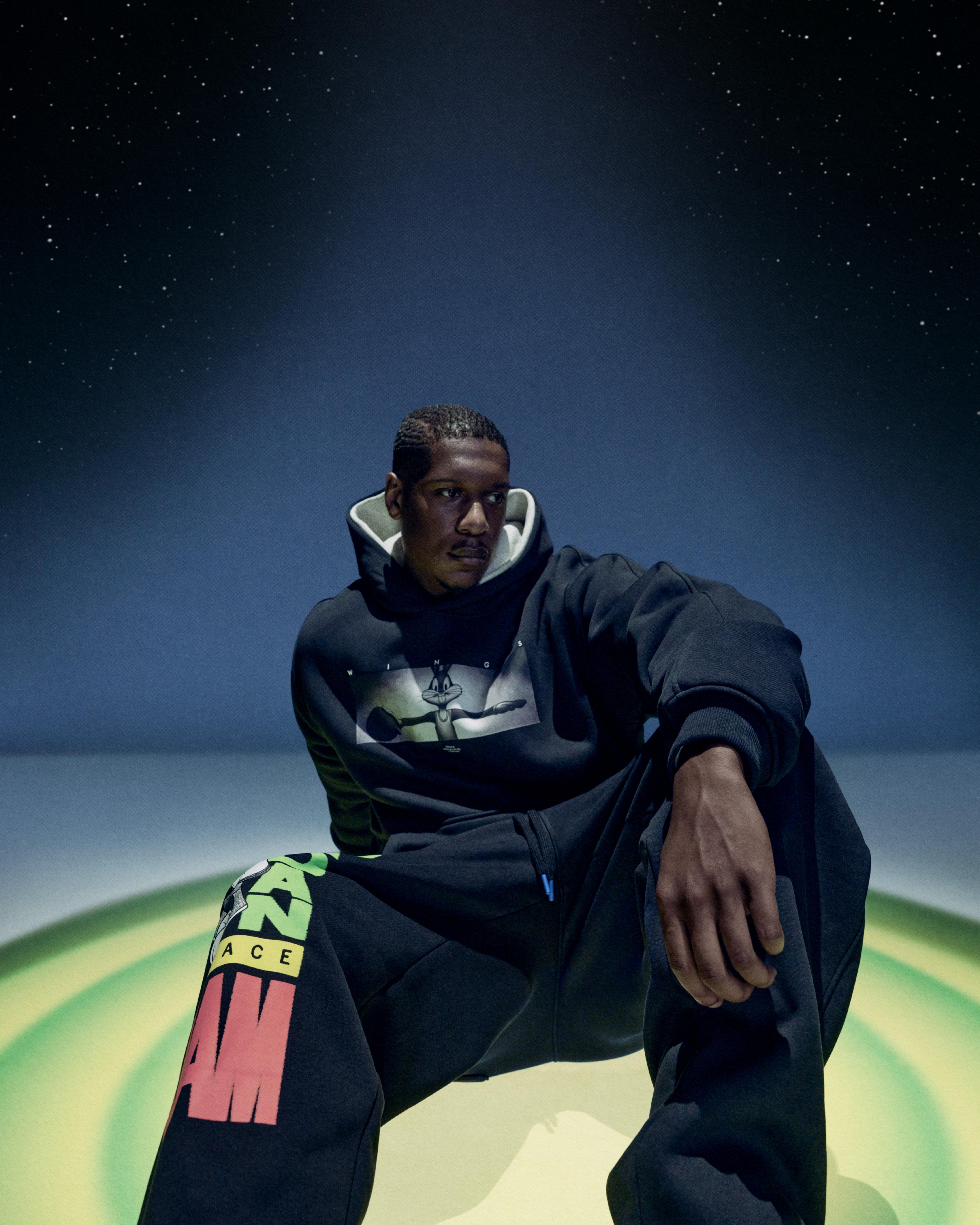 Person seated in a black Space Jam hoodie featuring Bugs Bunny with black sweatpants displaying colorful lettering against a starry blue background.