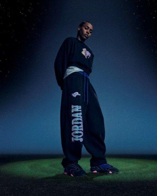 Person wearing an oversized black sweatshirt, black Jordan sweatpants with colorful graphics and black-and-pink sneakers against a starry blue background.