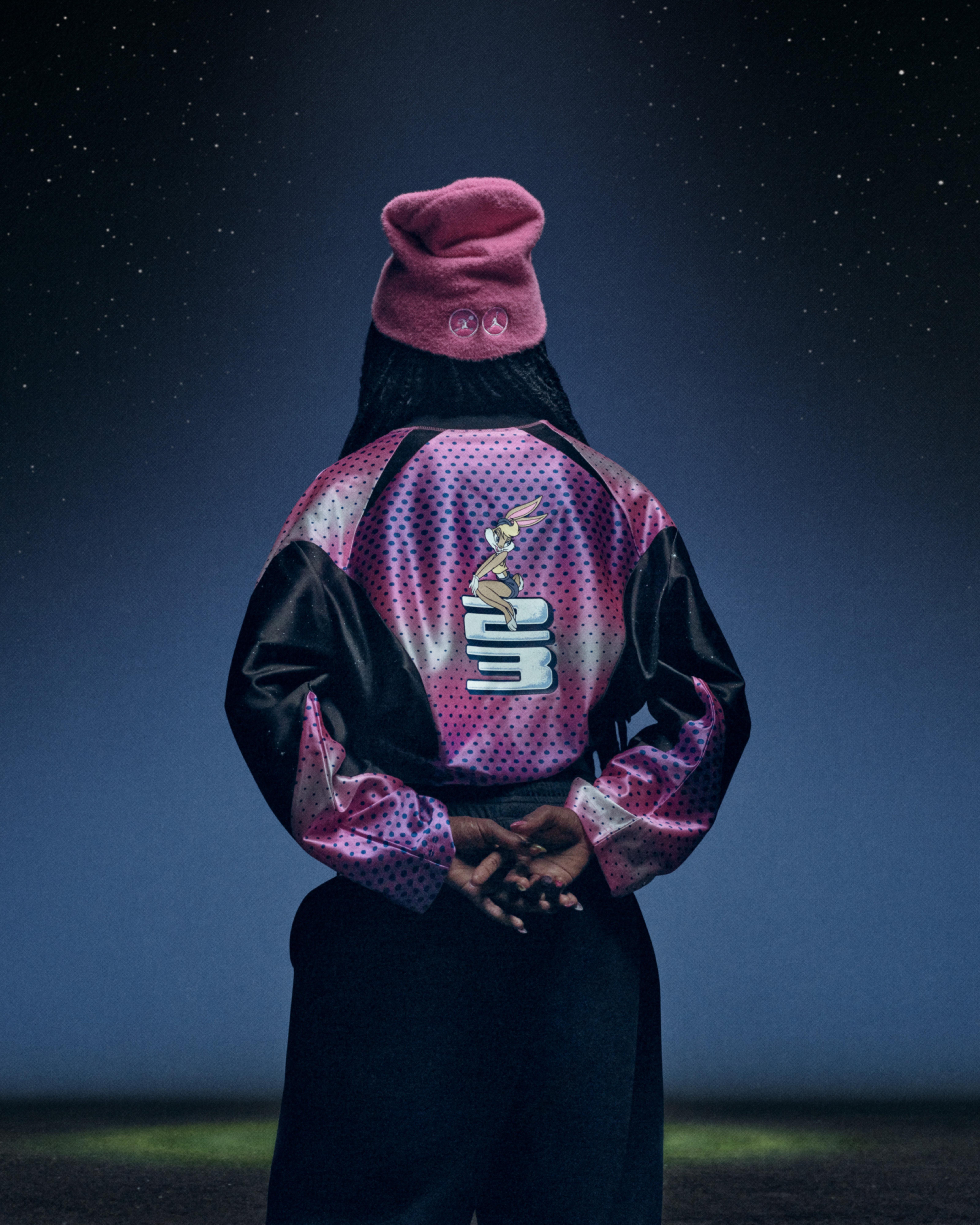Back view of a person wearing a pink-and-black perforated Jordan jacket featuring Lola Bunny and large Jordan lettering, paired with a pink fleece hat.