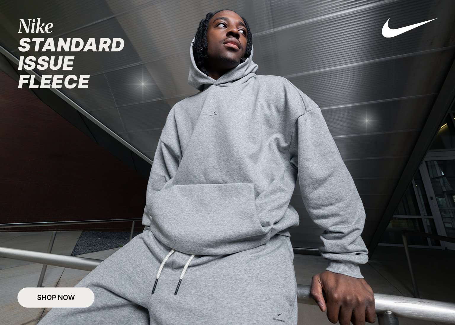 Man wearing Nike Standard Issue Fleece grey hooded sweatshirts and sweatpants, with a white Nike checkmark logo in the top right, and SHOP NOW in a white button.