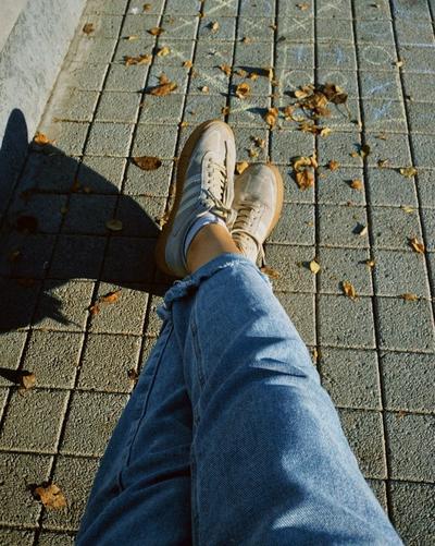 Top-down view of beige adidas Samba sneakers worn with light-wash jeans on a paved sidewalk with scattered leaves.
