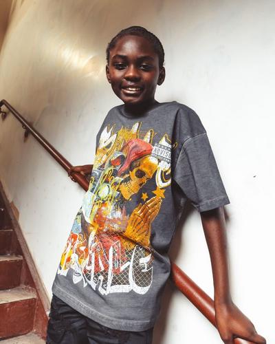 A smiling young boy leans against a wooden handrail on a staircase, wearing a charcoal grey oversized t-shirt featuring a large graphic of a praying skeleton in a red hood.