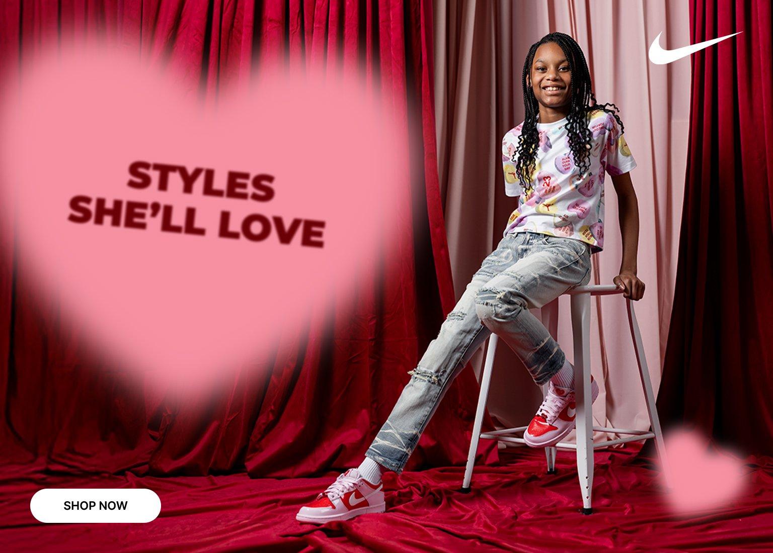  This promotional graphic for Nike features a young girl sitting on a white stool against a dramatic backdrop of red velvet curtains. She is wearing a colorful graphic t-shirt, light-wash distressed jeans, and red and white Nike sneakers. The image includes a large, soft pink heart containing the text "STYLES SHE'LL LOVE" in a bold red font.