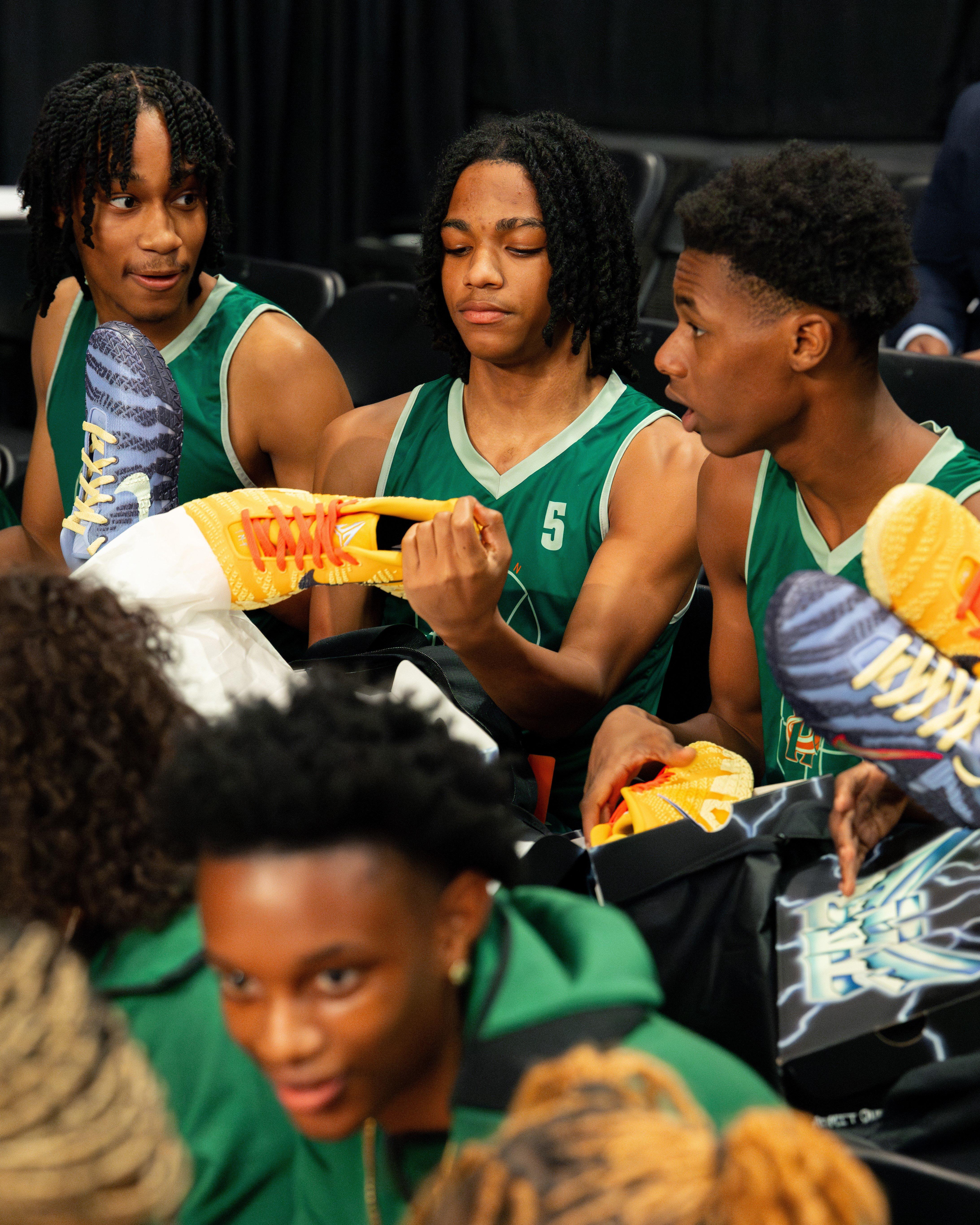 A group of high school basketball players in green jerseys are sitting in arena seating, examining brightly colored Nike basketball shoes in their hands.
