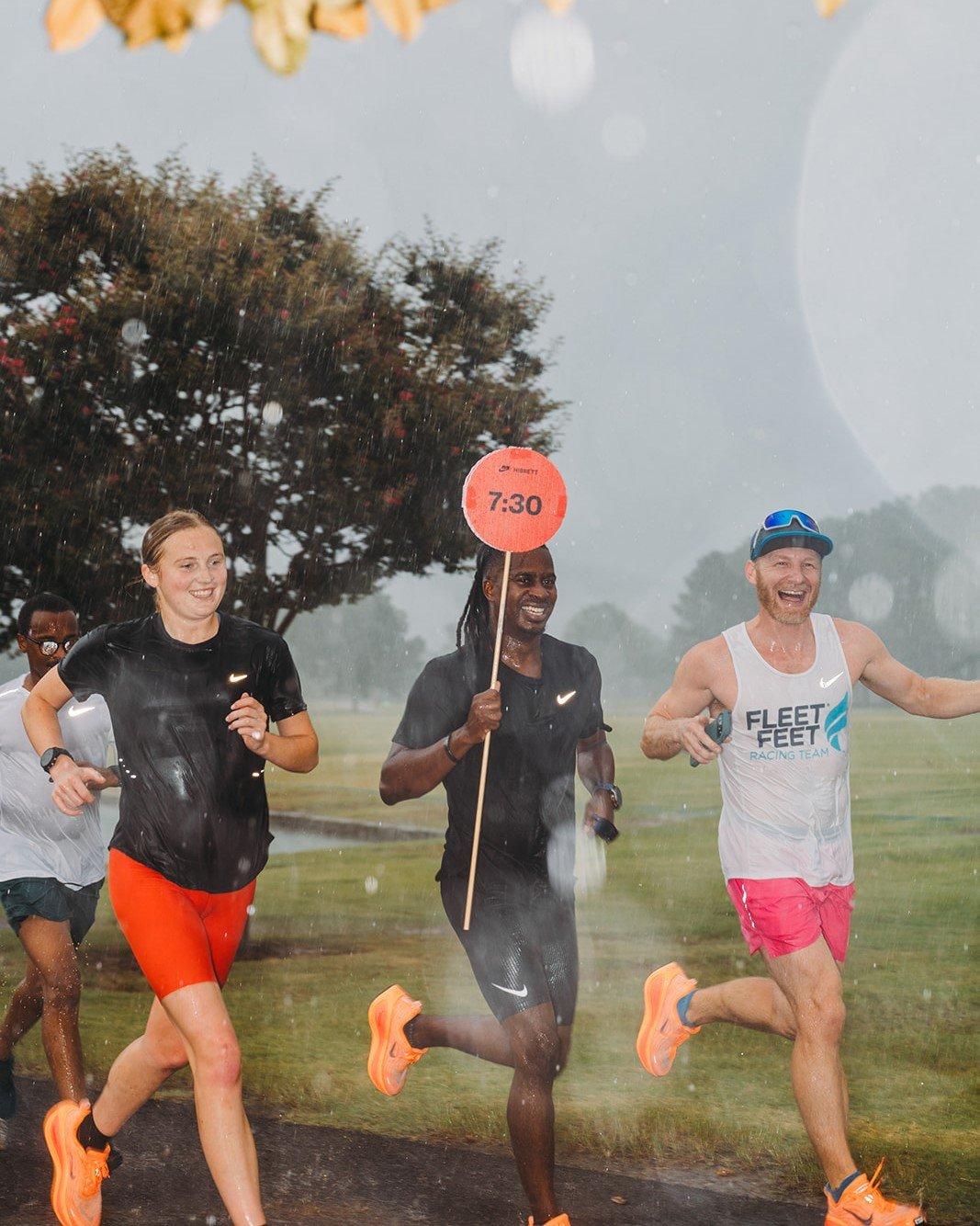 Three people are running and laughing in the rain on a paved path. The person in the middle is holding an orange sign on a stick that says "7:30". The person on the far right is wearing a tank top that says "FLEET FEET RACE TEAM." The background is a grassy area with a tree.