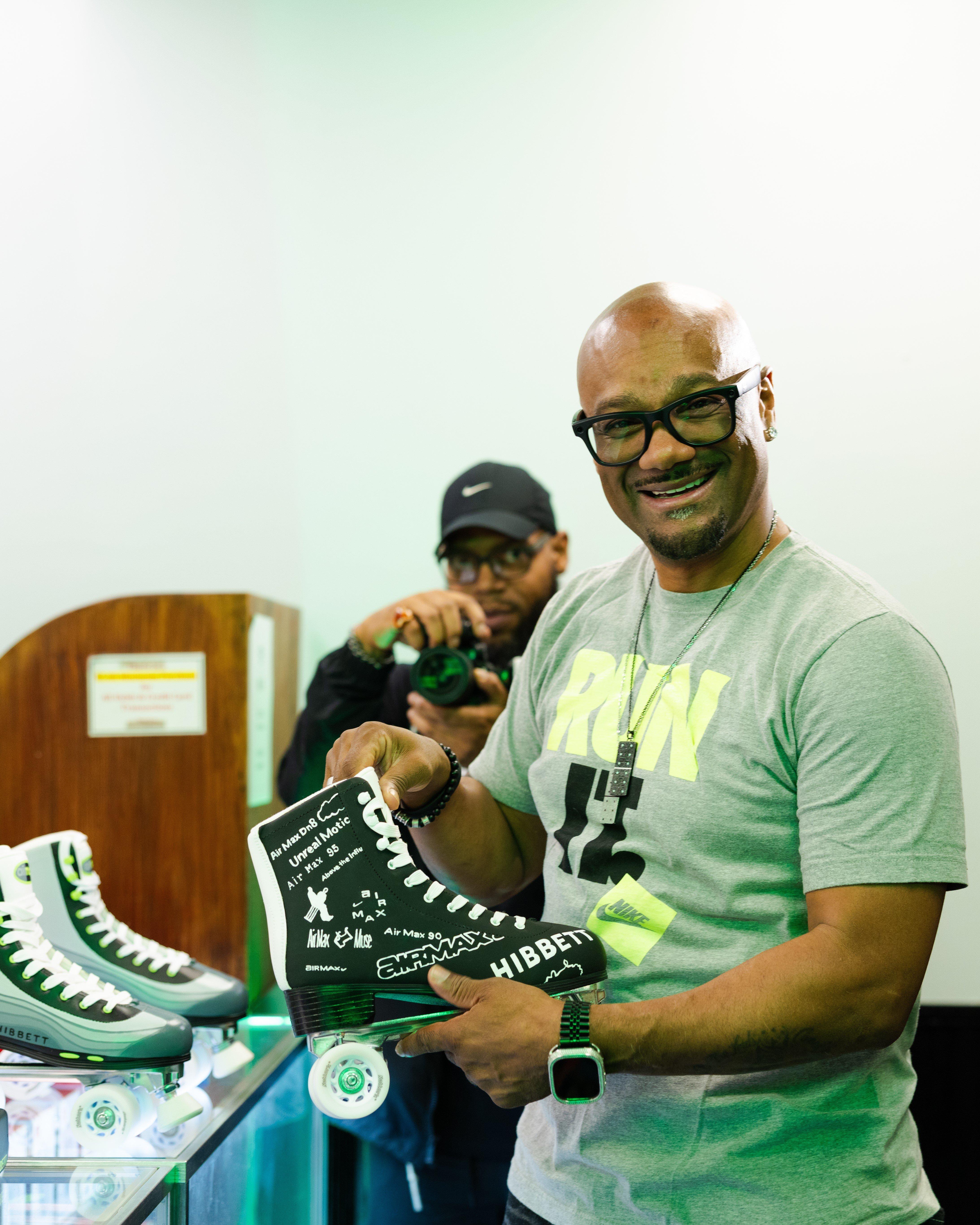 Media personality Big Tigger, wearing glasses and a grey t-shirt, smiles while holding up a custom black roller skate. The skate features white "Hibbett" and "Air Max" branding, while a photographer captures the moment in the background.