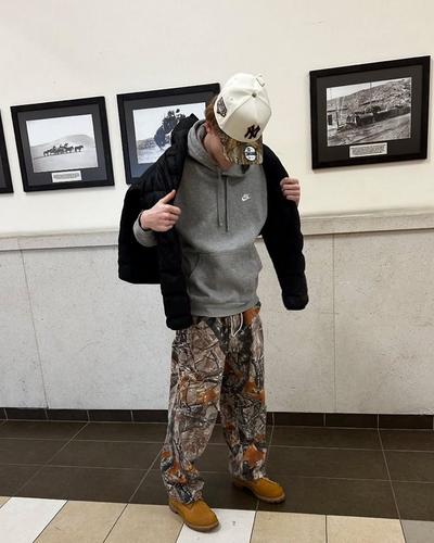 A young man poses indoors wearing wheat Timberland boots, camo print pants, a gray Nike hoodie, and a white New York Yankees cap with patches. He is adjusting a black puffer jacket over his shoulders while looking down. The setting features framed black-and-white historical photos on the wall.