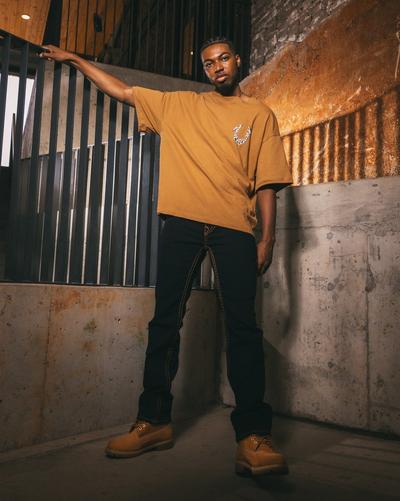 A full-body shot of a man standing in a dimly lit, industrial-style building. He is wearing a mustard-colored oversized t-shirt, black pants with visible yellow stitching, and brown work boots. He is holding onto a metal railing.
