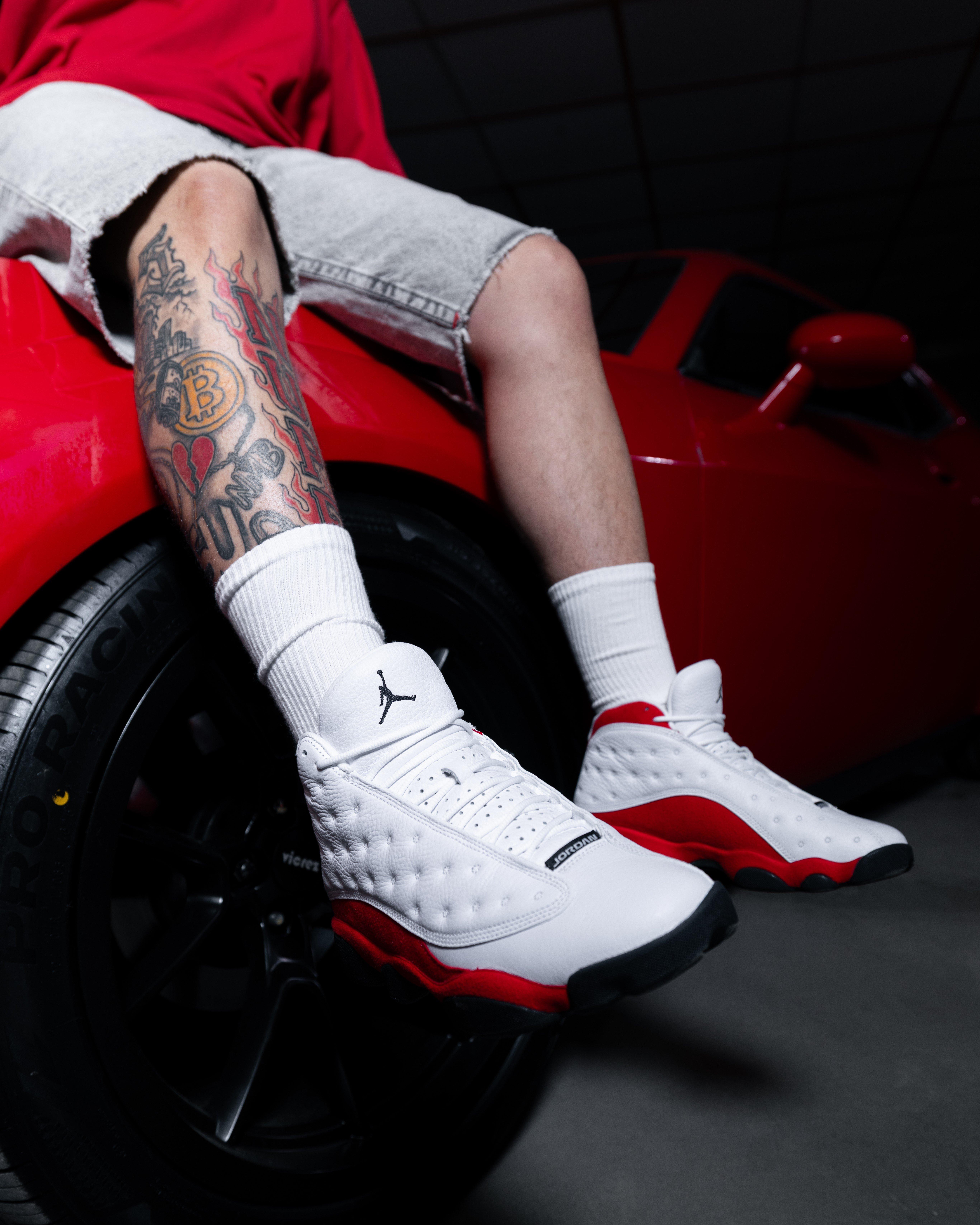 Model sitting on a red sports car wearing white and red Air Jordan 13 sneakers with white crew socks and denim shorts