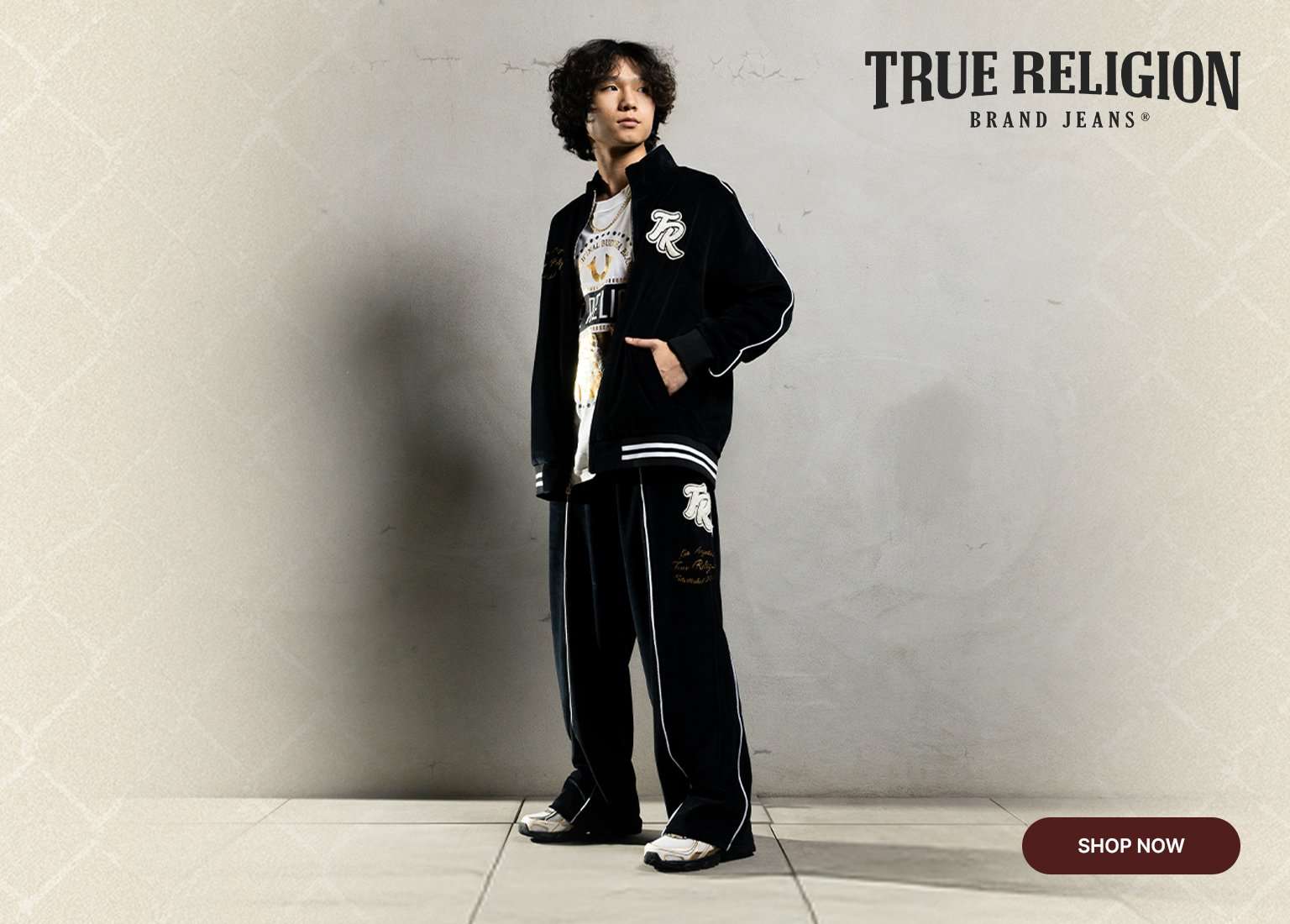 True Religion Brand Jeans featuring a model in a black velour tracksuit with white piping. Shop Now button.