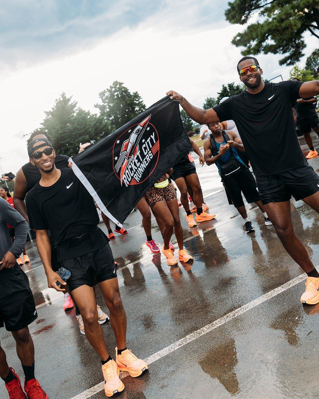 Two men stand smiling at the camera on a wet, paved surface. The man on the left is wearing sunglasses and holding one end of a black flag. The man on the right, also wearing sunglasses, holds the other end. The flag has a logo with "ROCKET CITY RUNNERS" on it. Other people are in the background.
