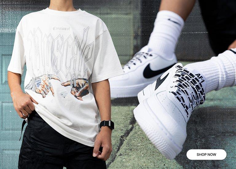 A promotional retail banner featuring an Upscale graphic tee with a "Creation of Adam" inspired hand illustration. On the right, a close-up shows white Nike Air Force 1 Low sneakers with branded laces and white socks. A "Shop Now" button is located in the bottom right corner.