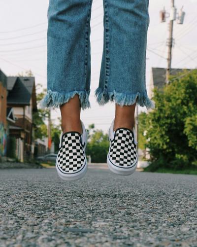 Close-up of black-and-white checkered Vans slip-on sneakers worn with frayed hem jeans on a paved street.