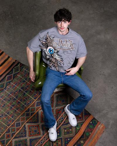 A high-angle shot of a man with curly brown hair, blue eyes, and wearing a gray Von Dutch t-shirt with a large eye graphic, blue jeans, and white sneakers. He is seated on a green chair on top of a multi-colored patterned rug.