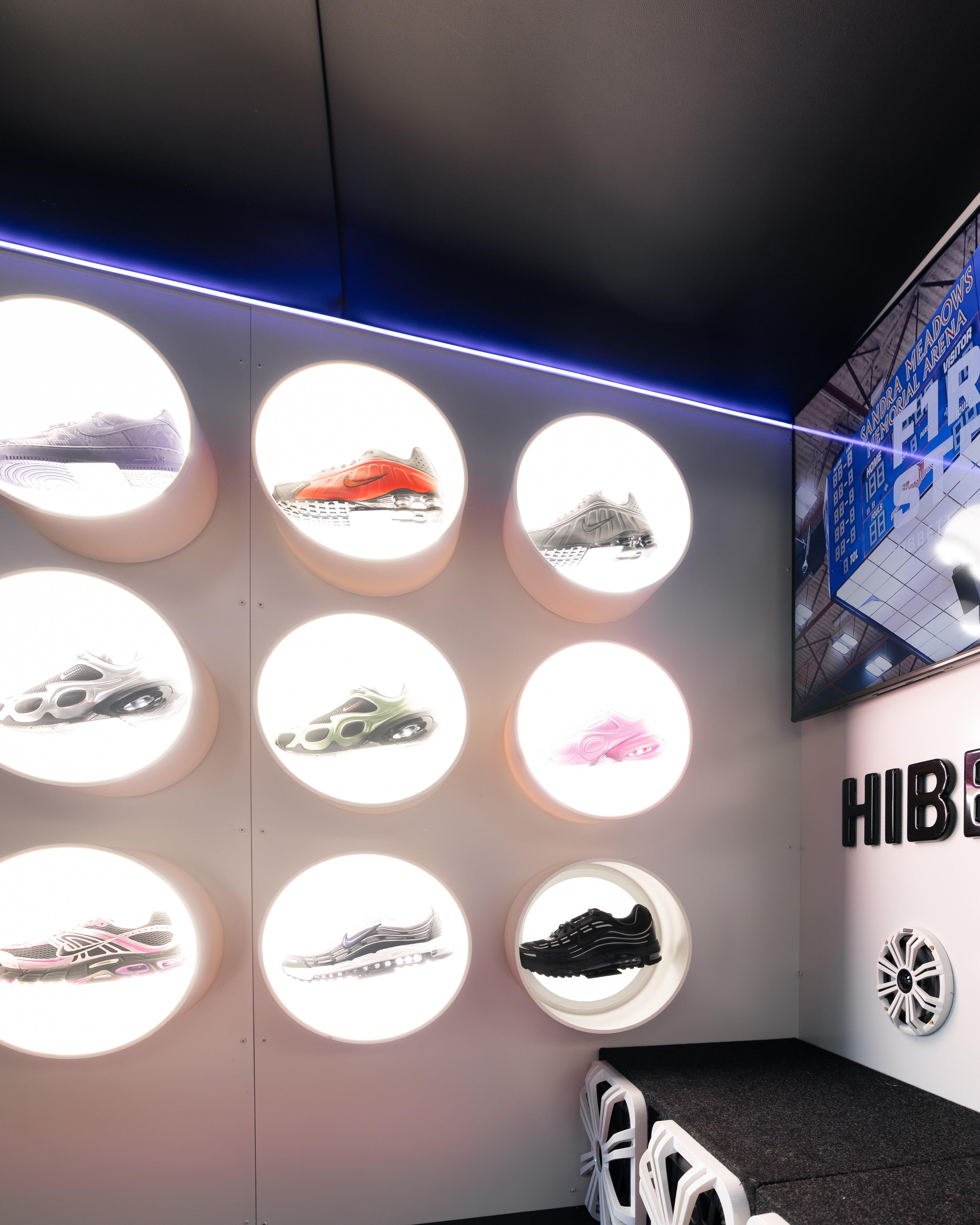 A modern display wall with multiple circular, illuminated shelves featuring various Nike footwear. A large screen to the right shows event information.
