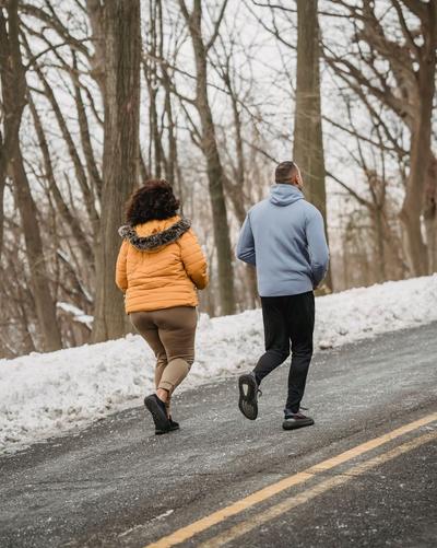 Two people jogging on a snow-lined road through a wooded area, wearing cold-weather jackets, athletic pants and running shoes.